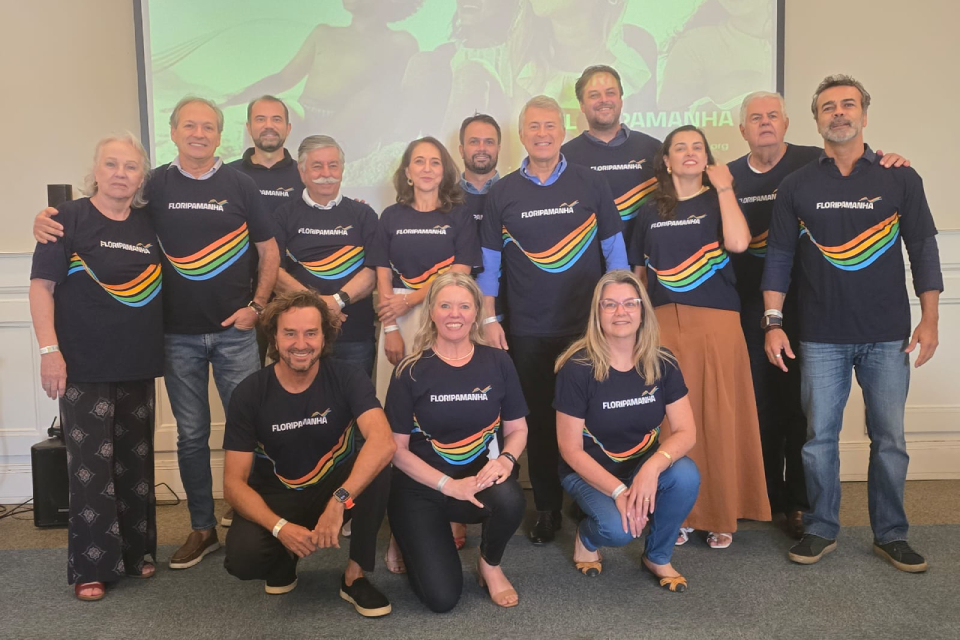 Fotografia de grupo com treze adultos usando camisetas pretas “FloripAmanhã”, posando sorridentes em ambiente interno, diante de tela projetada.