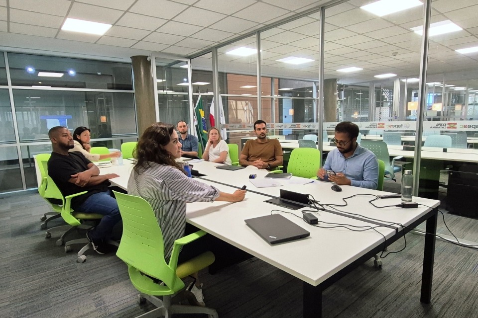 Fotografia de reunião em sala corporativa envidraçada, com sete pessoas sentadas à mesa branca, usando notebooks e cadernos, em cadeiras verdes, em clima de discussão e atenção coletiva.
