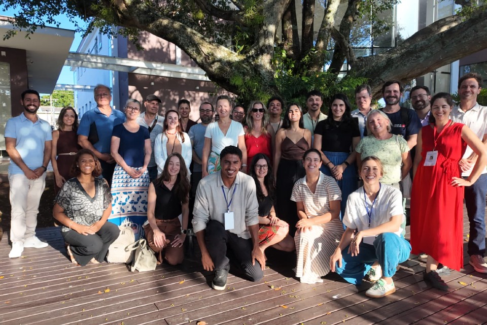 Fotografia de cerca de vinte e cinco adultos posando sorridentes sob uma grande árvore, em área externa com deck de madeira, diante de um prédio moderno. Alguns estão agachados na frente e outros em pé atrás, em clima descontraído.