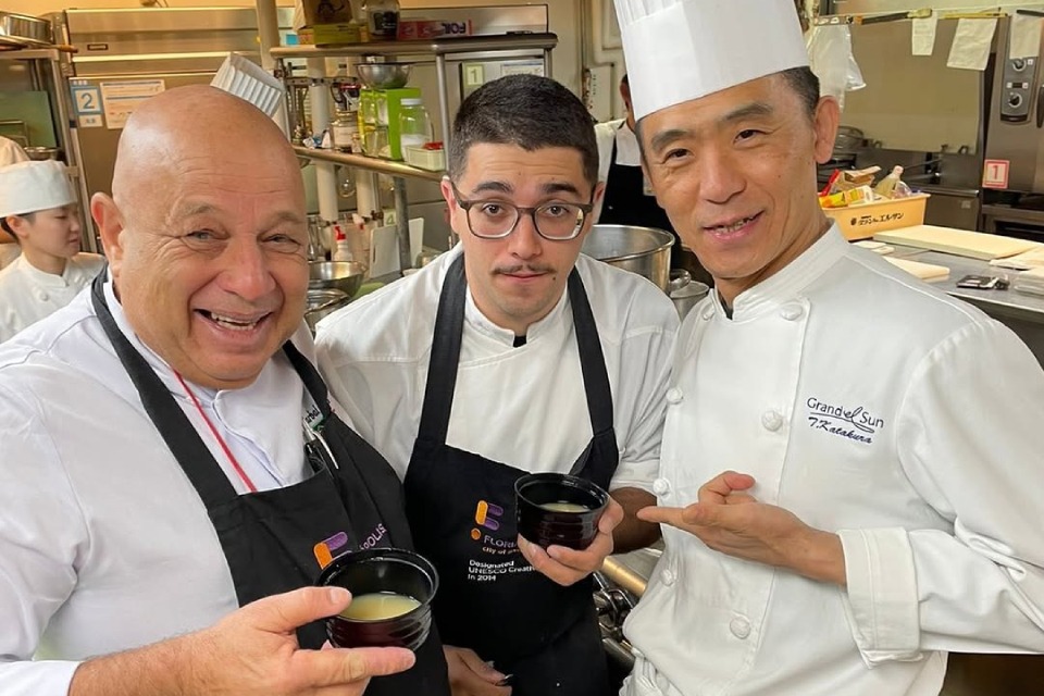 Fotografia em cozinha profissional: três chefs posam sorrindo, segurando pequenas tigelas com caldo; um careca de avental preto, um jovem de óculos ao centro e um chef asiático de dólmã branca apontando para a tigela.