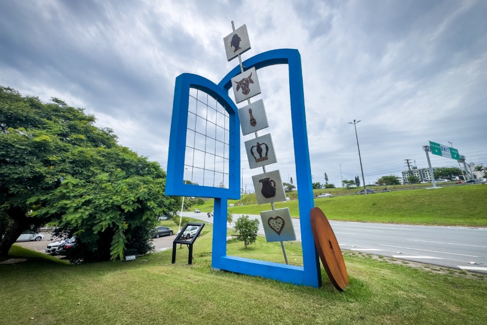 Monumento urbano ao ar livre, com estrutura azul em forma de arco e placas verticais com símbolos artesanais, instalado em área gramada à beira de uma avenida sob céu nublado.