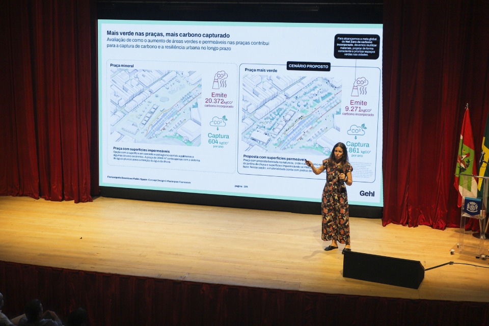 Fotografia de palestra em auditório: mulher de vestido estampado apresenta no palco diante de telão sobre áreas verdes, captura de carbono e resiliência urbana.