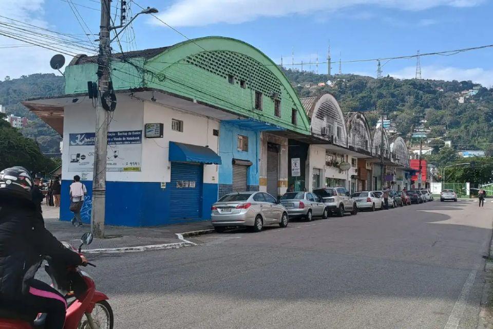 Fotografia de uma rua na antiga rodoviária de Florianópolis, SC. À esquerda, aparece uma pessoa de moto usando capacete, enquanto a rua segue à direita com diversos carros estacionados paralelamente ao longo de um prédio baixo e antigo, pintado nas cores azul e verde. O edifício possui telhados curvos e portas fechadas, aparentando ser um antigo complexo comercial. Ao fundo, há uma colina verde com casas e antenas, sob um céu parcialmente nublado.