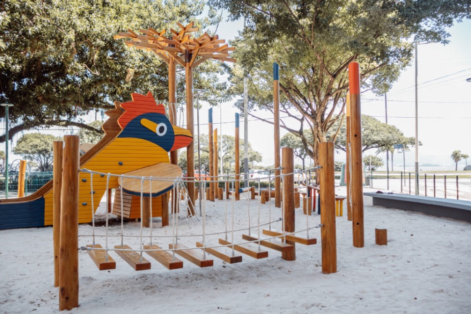 Fotografia de brinquedo de parque em forma de pássaro amarelo e azul, com escorregador, sobre areia e sob árvores.