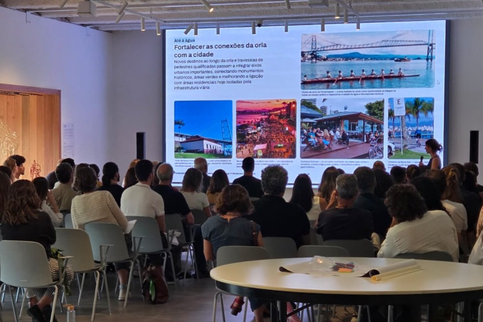 Fotografia de auditório com dezenas de pessoas sentadas diante de um telão. À direita, uma mulher acompanha a apresentação, que traz o título “Até a água — Fortalecer as conexões da orla com a cidade” e imagens de ponte, rua movimentada e espaço público à beira-mar.