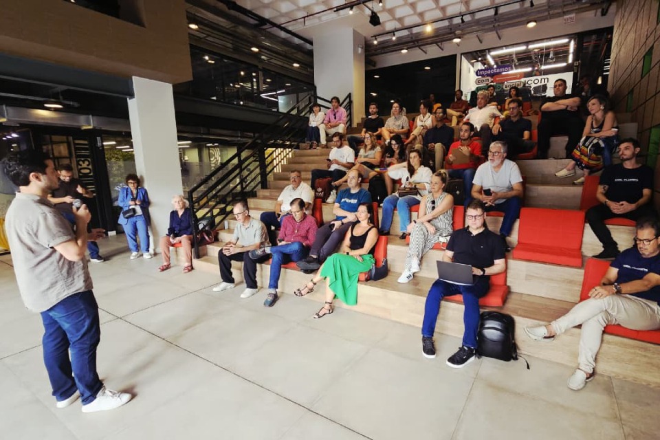 Fotografia de palestra em auditório moderno, com um homem falando ao microfone para dezenas de pessoas sentadas em arquibancada.
