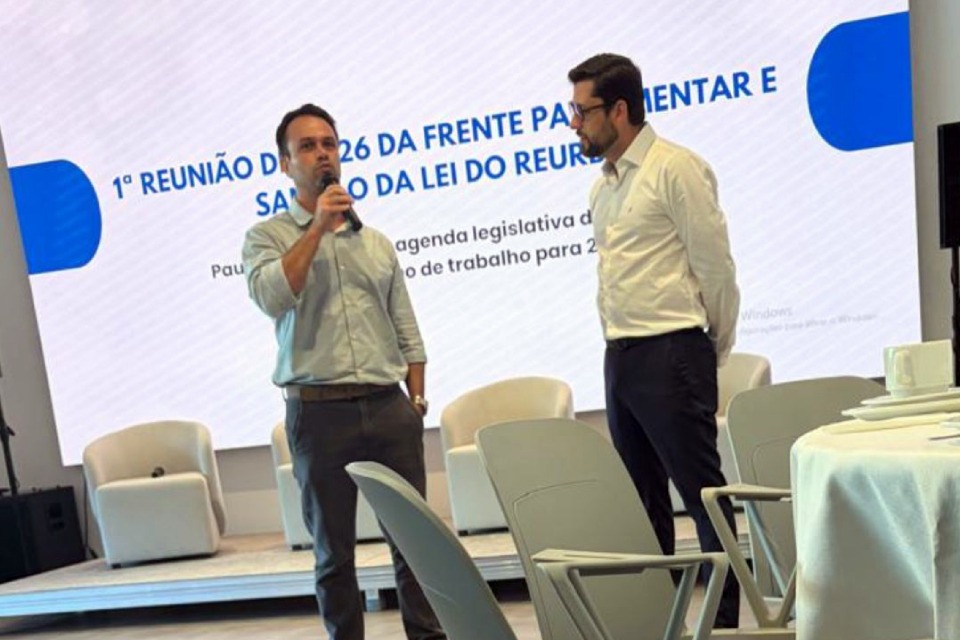 Fotografia de palco com dois homens de camisa social diante de telão institucional; um fala ao microfone enquanto o outro o observa. Ao fundo, aparece o texto: “Primeira reunião de dois mil e vinte e seis da Frente Parlamentar e sanção da lei do Reurb.”