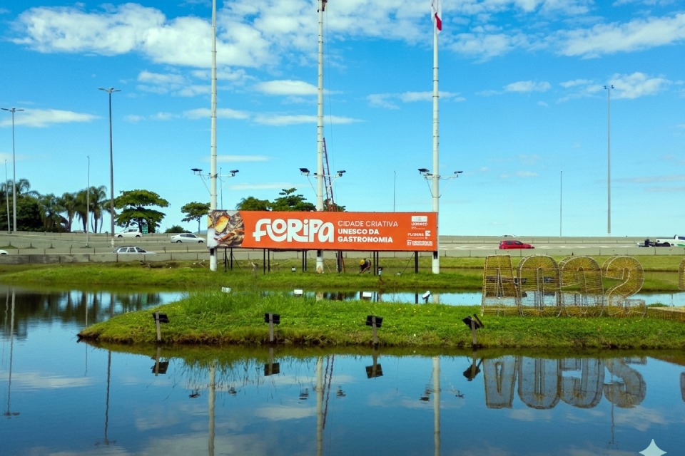 Fotografia de lago com reflexo do céu e de um gramado central, onde há um painel laranja com o texto “FLORIPA CIDADE CRIATIVA UNESCO DA GASTRONOMIA” e, à direita, a estrutura “dois mil e vinte e dois”. Ao fundo, aparecem pista com carros, postes altos e céu azul com nuvens.